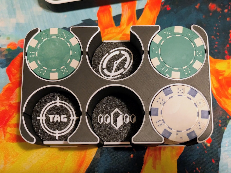 May include: A black plastic chip holder with six circular compartments, each holding a game chip. The chips are teal, white, and black, with various designs including a target, a compass, and the word "TAG".