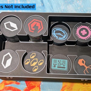 May include: Black game token organizer with multiple compartments, holding stacks of circular tokens. The tokens feature various geometric and abstract designs in white, red, blue, yellow, and orange. Text at the top reads "Tokens Not Included".