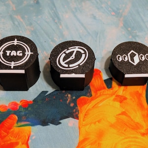 May include: Three black, cylindrical game tokens with white graphic designs. One token has a target with the word "TAG", another has a clock-like design, and the third has a geometric pattern. Each token has a white rectangular base.
