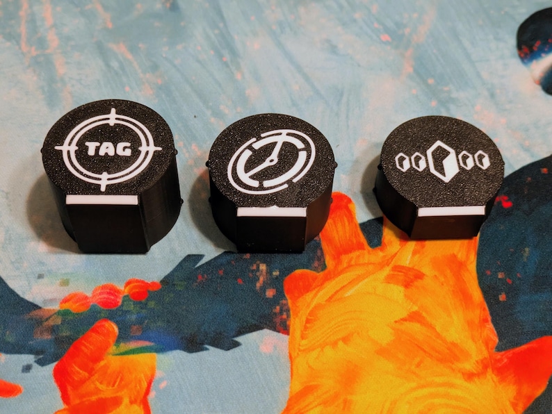 May include: Three black, cylindrical game tokens with white graphic designs. One token has a target with the word "TAG", another has a clock-like design, and the third has a geometric pattern. Each token has a white rectangular base.
