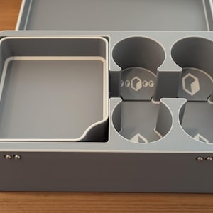 May include: A gray plastic organizer with a white trim, featuring compartments for storage. The organizer has a square tray, four circular cup holders, and a rectangular compartment. The lid is open, revealing the interior.