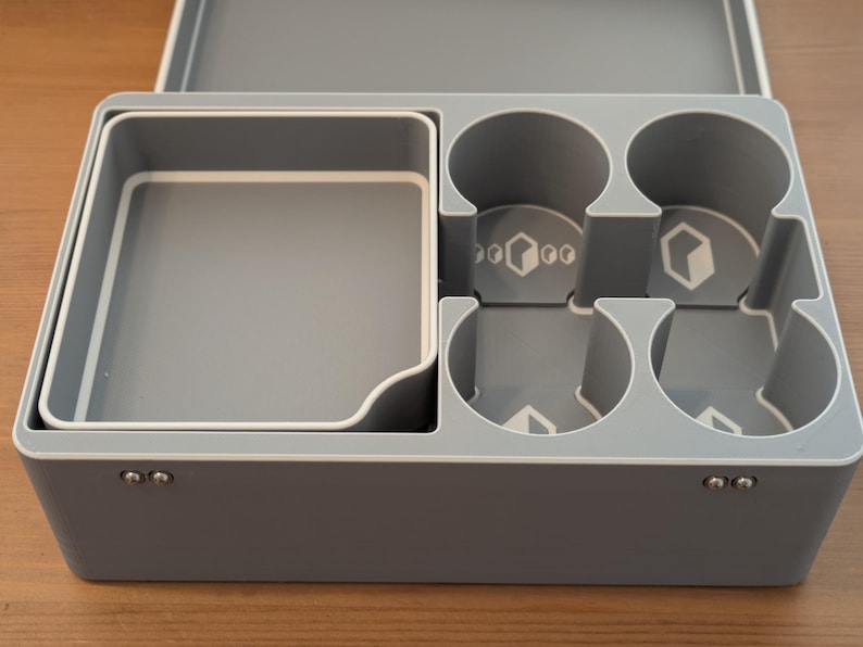 May include: A gray plastic organizer with a white trim, featuring compartments for storage. The organizer has a square tray, four circular cup holders, and a rectangular compartment. The lid is open, revealing the interior.
