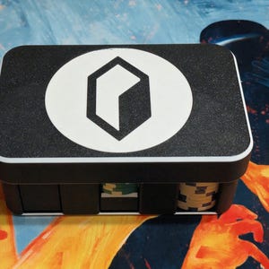 May include: A black rectangular box with a white geometric logo on the lid. The box is filled with poker chips. The lid has a textured surface and a white border. The logo is a white circle with a black hexagon inside.
