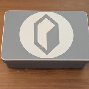 May include: A rectangular, light gray box with a white circular emblem featuring a gray geometric design. The box is sitting on a wooden surface.