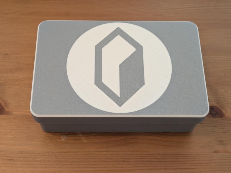 May include: A rectangular, light gray box with a white circular emblem featuring a gray geometric design. The box is sitting on a wooden surface.