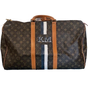 May include: A brown and tan Louis Vuitton travel bag with a white and blue striped design and the initials "K.M." embroidered on the front.