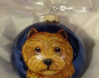 May include: A hand-painted glass bauble featuring a brown and white dog with a blue background. The dog has a fluffy coat and is looking directly at the viewer.