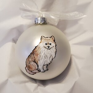 May include: A white glass Christmas ornament with a hand-painted image of a small, fluffy, brown and white dog. The dog is sitting and looking at the viewer.