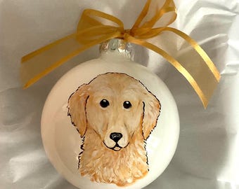 Puede incluir: Una bola de Navidad blanca con un retrato pintado a mano de un perro golden retriever. El perro tiene un pelaje marrón claro y ojos negros. Una cinta dorada está atada en la parte superior. Este adorno festivo es perfecto para los amantes de los perros.
