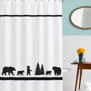 May include: A white shower curtain featuring a horizontal black stripe near the top and bottom. The lower section displays a black silhouette pattern of bears and pine trees. The curtain hangs from a silver rod with metal rings at the top.