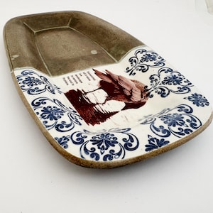 May include: A ceramic decorative tray with a brown and white color scheme. The tray features a brown upper section and a white lower section with blue floral patterns. A bird illustration and text are in the center.