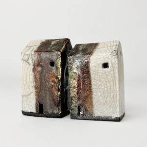 Op de afbeelding: Twee kleine keramische huisjes met een witte glazuur en bruine en gouden accenten. De huisjes zijn aan elkaar verbonden en hebben kleine openingen die lijken op ramen.