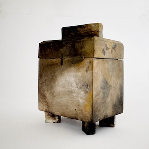 May include: A rectangular box with a lid and small feet, showing signs of age. The box has a textured surface with brown, grey, and yellow hues. The lid features a raised rectangular section. The box is likely for storage, with a rustic aesthetic.