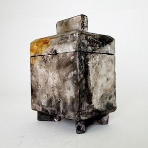 May include: A square ceramic lidded box with a textured, mottled finish in shades of charcoal, gray, and white. The container features a rectangular handle on the lid and stands on four short, block-like feet, displaying a weathered appearance.