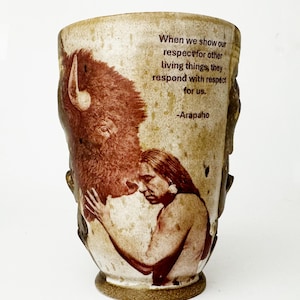 Puede incluir: Taza de cerámica con fondo beige e imagen marrón de un bisonte y una persona nativa americana. La taza tiene la cita: "Cuando mostramos nuestro respeto por otros seres vivos, responden con respeto por nosotros." -Arapaho.