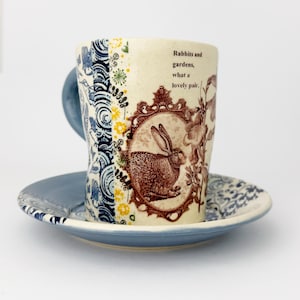 Puede incluir: Un juego de taza y platillo de cerámica con un patrón floral azul y blanco. La taza tiene una ilustración de conejo marrón con el texto "Rabbits and gardens, what a lovely pair."