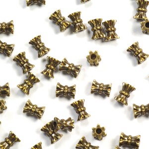 May include: A scattering of antique gold-toned metal beads with a bow design. The beads are small and have a decorative, ornate look.