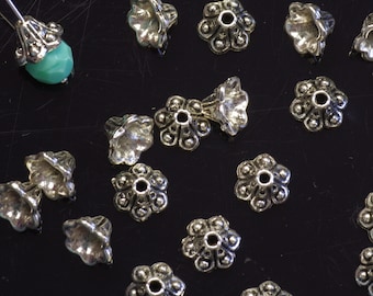 Op de afbeelding: Een verzameling zilverkleurige metalen kralencaps met een bloemvormig ontwerp. De caps hebben een klein gaatje in het midden om kralen door te rijgen.