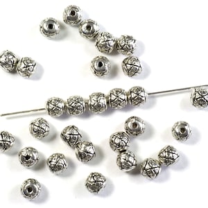May include: A scattering of silver metal beads with a geometric pattern. The beads are arranged in a line on a white surface.
