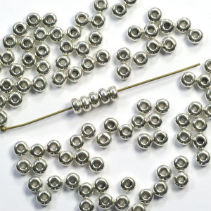 Op de afbeelding: Een grote hoeveelheid zilverkleurige metalen kralen, waarvan sommige op een dun draad geregen zijn. De kralen zijn rond met een gat in het midden.