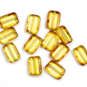 Könnte beinhalten: Eine Sammlung rechteckiger, bernsteinfarbener Glasperlen mit durchscheinendem Aussehen. Jede Perle hat eine hellgelbe Mitte und dunklere Bernsteinkanten, mit drei parallelen Linien, die durch die Mitte verlaufen. Die Perlen sind auf einer weißen Oberfläche verstreut.