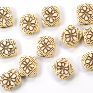 May include: A collection of twelve off-white square beads with a brown floral design. The beads are arranged in a scattered pattern on a white surface.