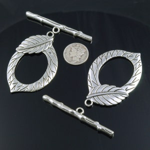 Könnte beinhalten: Zwei silberfarbene Metall-Toggle-Verschlüsse mit Blattmotiven. Jeder Verschluss hat eine runde Öffnung und ein dekoratives Blattmotiv. Die Verschlüsse sind etwa 5 cm lang.