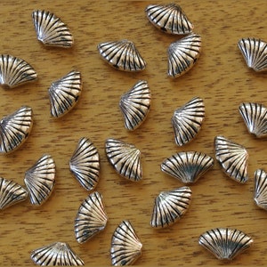 May include: A collection of silver-toned metal shell-shaped charms, arranged on a wooden surface. The charms are all the same shape, but vary in size and orientation.