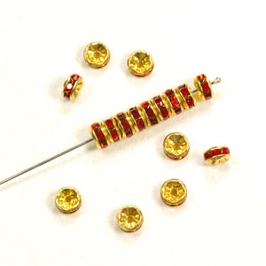 May include: A close-up of a string of gold-toned beads with red crystal accents. The beads are strung on a thin wire and are scattered on a white surface.