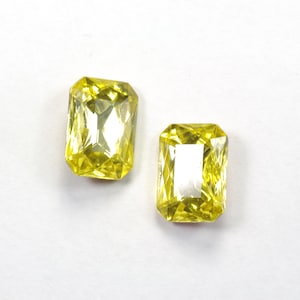 May include: Two yellow emerald cut gemstones on a white background.