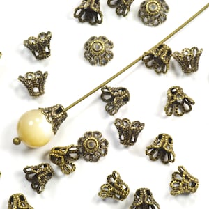 May include: A collection of antique gold-toned metal bead caps with a decorative, scalloped design. The caps are scattered on a white surface, with one cap on a white bead.