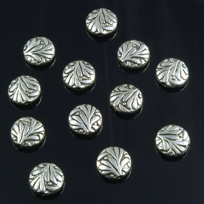 Pode incluir: Doze contas de metal prateadas com um design decorativo de folhas. As contas s&atilde;o redondas e t&ecirc;m um acabamento liso e brilhante.