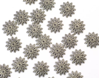 May include: A collection of silver-toned metal snowflake-shaped embellishments with a decorative design. The embellishments are scattered on a white background.