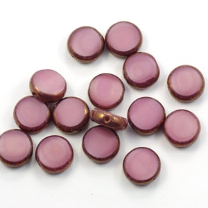 May include: A collection of round, flat beads with a light purple center and a dark purple rim with gold accents. The beads are scattered on a white surface, showcasing their design and color variations.