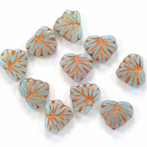 May include: A collection of ten light blue glass beads shaped like leaves with a copper-colored design. The beads are scattered on a white surface.