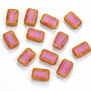 May include: A collection of rectangular beads with a pink center and a brown border. The beads are scattered on a white surface. The pink center has a striped pattern.