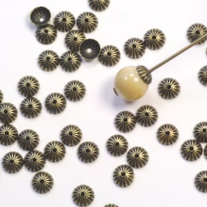 May include: A collection of antique bronze-colored bead caps with a starburst design. A light-colored bead is attached to a long, thin metal rod. The bead caps are scattered on a white surface, ideal for jewelry making.