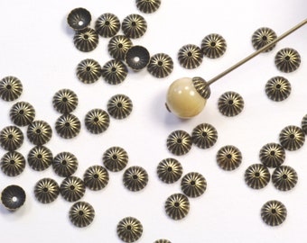 May include: A collection of antique bronze-colored bead caps with a starburst design. A light-colored bead is attached to a long, thin metal rod. The bead caps are scattered on a white surface, ideal for jewelry making.