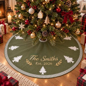 May include: A round, olive green knit Christmas tree skirt featuring a white striped border and a repeating pattern of white pine trees. The center is embroidered with the gold text "The Smiths" above "Est. 2026" and two gold leaf sprigs.