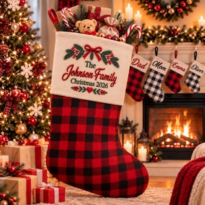 May include: A red and black buffalo check Christmas stocking with a cream-coloured cuff featuring embroidered green pine branches, red berries, and the text The Johnson Family Christmas 2026. The stocking is filled with a teddy bear and candy canes.