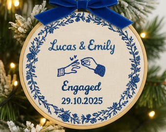 May include: A round, cream-colored embroidered ornament with the names "Lucas & Emily" and the word "Engaged" in blue script. The ornament features a blue floral border and a blue velvet bow at the top. The date "29.10.2025" is also included.