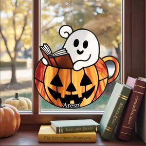 Puede incluir: Decoración de Halloween de vidriera que muestra un fantasma sonriente leyendo un libro dentro de una calabaza naranja. La calabaza tiene rasgos negros recortados y la palabra "Aresn". Libros y calabazas están sobre una superficie de madera.