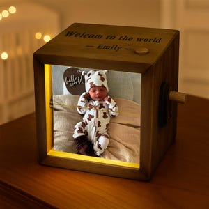 May include: A wooden photo cube with a lit interior, displaying a baby in a bear-patterned outfit. The top of the cube reads "Welcome to the world - Emily -". A small "hello world" sign is also visible.