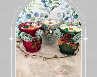 May include: Three decorative candles in red, silver, and green containers. Each candle is adorned with pinecones, berries, and faux evergreen branches. The candles are arranged on a marble surface with a string of small bells.