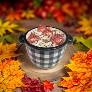 May include: A black and white checkered bowl filled with whipped cream, decorative sprinkles, and red ornament-shaped chocolates. The bowl sits on a wooden surface surrounded by colorful autumn leaves, creating a festive fall theme.