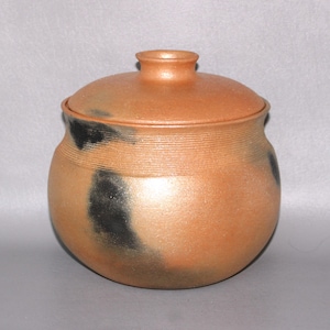 May include: A round, terracotta-colored ceramic jar with a matching lid. The vessel features a textured surface with horizontal grooves near the neck and irregular, dark charcoal-black smoke-like markings across the warm orange-brown body.