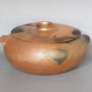 May include: A handmade, round ceramic pot with a lid. The pot is a warm, earthy orange color with dark gray and black speckled accents. It has two small handles and a knob on the lid.