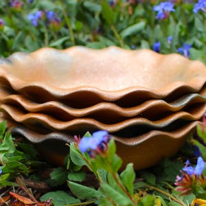 May include: A set of four brown ceramic bowls with a wavy edge. The bowls are stacked on top of each other and are sitting on a bed of green leaves and blue flowers.