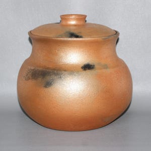 May include: A round, terracotta-colored ceramic jar with a matching lid. The surface features a matte, textured finish with irregular, dark charcoal-colored smoke-like markings scattered across the body and the lid, creating a mottled appearance.