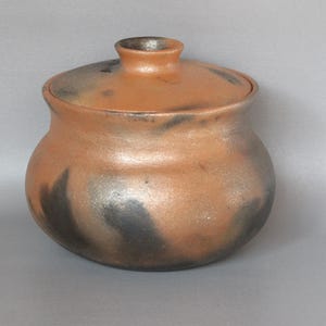 May include: A handcrafted ceramic jar with a lid. The jar is a warm orange-brown color with dark gray and black accents. The jar has a rounded body and a small, cylindrical neck. The lid fits snugly on top.